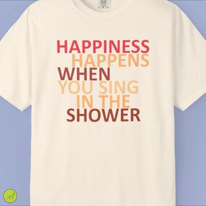 Op de afbeelding: Een crèmekleurig T-shirt met korte mouwen en de tekst "HAPPINESS HAPPENS WHEN YOU SING IN THE SHOWER" in een gestapelde lay-out. De letters gebruiken een verloop van rode, oranje en bruine tinten over de vijf regels tekst.