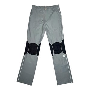 May include: Light gray pants featuring black circular knee patches and white piping accents along the legs. The pants include a button closure at the waist, belt loops, and a small white rectangular label on the lower left leg near the knee.