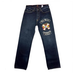 May include: Dark blue denim jeans featuring a Von Dutch logo patch on the left thigh. The patch includes a winged eyeball graphic surrounded by flames, with the text Von Dutch above and Compton California below in a gothic-style white font.