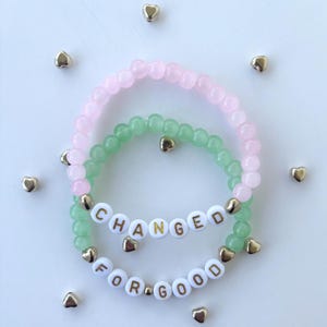 May include: Two beaded bracelets, one pink and one green, with the words "CHANGED FOR GOOD" spelled out in white letter beads. Gold heart-shaped beads are scattered around the bracelets.