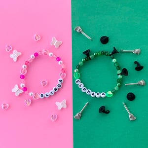 Może przedstawiać: Dwa bransoletki przyjaźni, jedna różowa z białymi koralikami i imieniem "Glinda" napisanym, druga zielona z zielonymi koralikami i imieniem "Elphaba" napisanym. Obie bransoletki mają zawieszki, jedna z motylami, a druga z miotłami i kapeluszami czarownic.