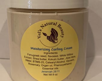 Op de afbeelding: Een doorzichtige plastic pot met een wit deksel, met een beige crème. Een geel etiket leest "Vel's Natural Beauty" en "Moisturizing Curling Cream". Het etiket vermeldt ingrediënten en het nettogewicht van 227g.