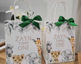 May include: Two white gift bags with green ribbon bows. The bags have a jungle animal theme with a giraffe, zebra, lion, and elephant. The bags are decorated with green leaves and the text "Zayn Turns One".