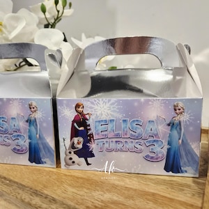 May include: Two silver metallic gable boxes featuring a printed graphic of characters from a winter-themed movie. The front of the boxes displays the text "ELISA TURNS 3" alongside illustrations of two female figures and a snowman on a light purple background.