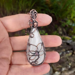 May include: A handcrafted copper wire pendant necklace featuring a teardrop-shaped, white stone. The stone is adorned with a copper wire dragonfly design and a small, pink gemstone at its center. The pendant hangs from a copper chain.