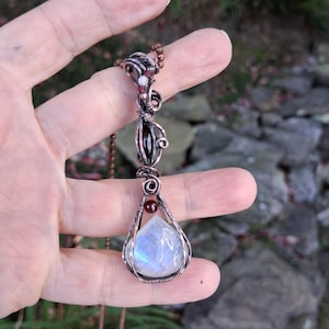 May include: A wire-wrapped pendant featuring a teardrop-shaped moonstone at the base, accented by a small dark red bead. The copper wire design extends upward, incorporating a dark oval stone and additional small beads, suspended on a chain.