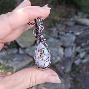 May include: A teardrop-shaped moonstone pendant wrapped in oxidized copper wire. The wirework features intricate swirls, small metal beads, and a tiny star-shaped charm. The pendant is suspended from a copper ball chain, showcasing a polished finish.