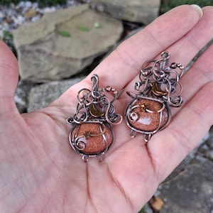 May include: Two pendants crafted from copper wire, each featuring a round, sparkling brown sandstone cabochon shaped like a cauldron. The wirework includes decorative swirls, a small crescent moon, and a tiny star accent on each piece.