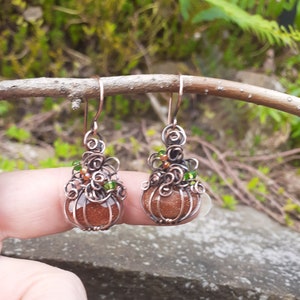 May include: A pair of handmade copper wire earrings. Each earring features a round, sparkling, orange-brown stone encased in a wire cage. The top of each earring is adorned with wire swirls and small green and orange beads.