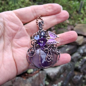 May include: A handcrafted pendant featuring a large, polished amethyst stone encased in copper wire. The pendant is adorned with purple flowers and a butterfly, with a copper chain. The design is intricate and detailed, showcasing a unique, artistic style.