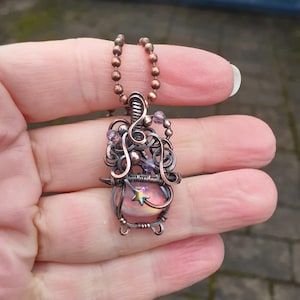 May include: A handcrafted copper wire pendant necklace. The pendant features a pink and iridescent stone with a star accent, surrounded by intricate wirework and purple beads. The necklace is suspended from a copper chain.