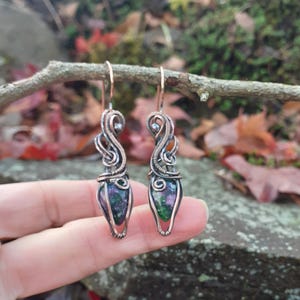 May include: A pair of handcrafted earrings featuring a teardrop-shaped, multi-colored stone wrapped in copper wire. The earrings have a unique, swirling design with a hook closure. The stones display shades of green, purple, and blue.