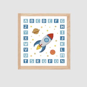 Puede incluir: Un bordado a punto de cruz enmarcado con un cohete, planetas y estrellas. El diseño espacial está rodeado por el alfabeto. El cohete es blanco con detalles azules y rojos. Los planetas son marrones y naranjas. El marco es de madera clara.