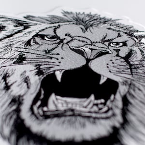 Op de afbeelding: Een close-up van een zwart-wit geborduurd leeuwenhoofd met een woeste uitdrukking. De leeuwenmanen zijn gedetailleerd en de tanden zijn zichtbaar.