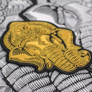 Könnte beinhalten: Ein gold gestickter Patch mit dem Kopf eines Elefanten mit aufwendigen Details. Der Patch ist auf schwarzem Hintergrund und hat einen weißen Rand.