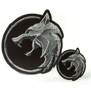 May include: Two embroidered patches featuring the Witcher logo. The larger patch is black with a silver wolf head, while the smaller patch is black with a silver wolf head.