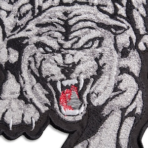Op de afbeelding: Een wit geborduurde tijgerpatch op een zwarte achtergrond. De tijger gromt met zijn bek open en zichtbare tanden. De ogen van de tijger zijn zwart en zijn vacht is gedetailleerd met wit en zilverdraad.