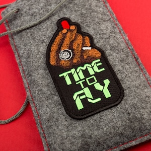 Peut inclure: Une pochette en feutre gris avec un patch brodé noir et vert qui dit "Time to Fly". Le patch représente une main brune faisant un signe de paix.
