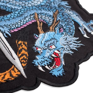 以下が含まれることがあります： 赤い目、白い歯、茶色の角を持つ、詳細な青いドラゴンの刺繍パッチ。 デザインには、白い剣とオレンジ色の虎の縞模様が黒い背景にあります。 パッチはユニークな波状の縁を持っています。