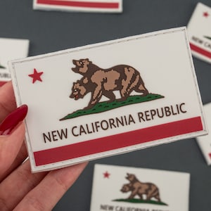 Könnte beinhalten: Ein weißer Patch mit einem roten Streifen am unteren Rand. Der Patch zeigt einen braunen Bären mit einem roten Stern darüber. Der Text "NEW CALIFORNIA REPUBLIC" ist in braunen Buchstaben gedruckt.