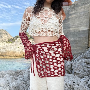 Puede incluir: Un top de ganchillo calado color crema con mangas largas y una falda envolvente de ganchillo roja a juego. El conjunto se combina con pantalones color crema. El fondo presenta una costa rocosa y cielo azul.