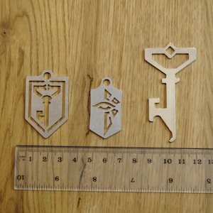 Pode incluir: Três chaveiros de metal com designs diferentes. O primeiro chaveiro é um escudo prateado com uma fechadura e uma chave dentro. O segundo chaveiro é um escudo prateado com um design de olho estilizado. O terceiro chaveiro é um abridor de garrafas prateado com um design de fechadura estilizado.