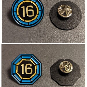 May include: Two enamel pins with the number 16 in a yellow circle on a blue background. The first pin is round and the second pin is octagonal. Both pins have a black backing with a metal pin.