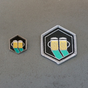 Könnte beinhalten: Zwei goldene Bierkrüge mit grünem und schwarzem Hintergrund auf einer grauen Oberfläche. Die Krüge sind silberfarben umrandet und haben einen weißen Schaum oben. Der Hintergrund ist ein schwarzes Sechseck mit silbernem Rand.