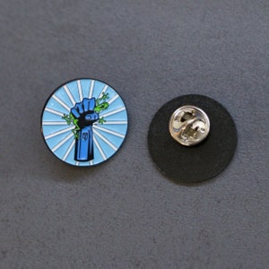 May include: A blue enamel pin with a raised fist and a green frog on top. The background is a blue and white striped design.