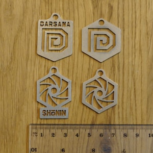 Pode incluir: Quatro pingentes de metal prateado com desenhos recortados. Dois pingentes são retangulares com a palavra "DARSANA" e um "D" estilizado. Os outros dois são hexagonais com a palavra "SHONIN" e um desenho em espiral. Uma régua está na parte inferior.