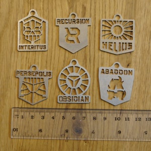 Pode incluir: Seis chaveiros de metal prateado com diferentes designs e texto. Os chaveiros apresentam as palavras "Interitus", "Recursion", "Helios", "Persepolis", "Obsidian" e "Abaddon".