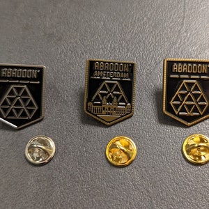Pode incluir: Três pinos de esmalte preto e dourado com o texto "ABADDON" e "ABADDON AMSTERDAM". Os pinos estão sobre uma superfície cinza com três costas de pinos de metal dourado.