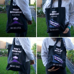 Könnte beinhalten: Vier Tragetaschen mit verschiedenen Sprachen darauf. Die Taschen sind schwarz mit weißem Text und einer lilafarbenen Grafik einer Flasche. Der Text auf den Taschen lautet "Don't push me! I have in my bag", "Nicht Schubsen! Ich habe im Beutel", und "Don't push me! I have in my bag".