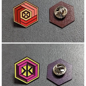 Könnte beinhalten: Zwei Emaille-Pins mit geometrischem Design. Der erste Pin ist rot, gelb und orange mit einer schwarzen Mitte. Der zweite Pin ist lila, gelb und pink mit einer schwarzen Mitte. Beide Pins haben eine silberne Rückseite mit einer Sicherheitsnadel.