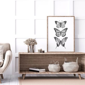 May include: A framed black and white print of three butterflies on a white background. The print is hanging on a white wall above a wooden shelf with two woven baskets and a book.