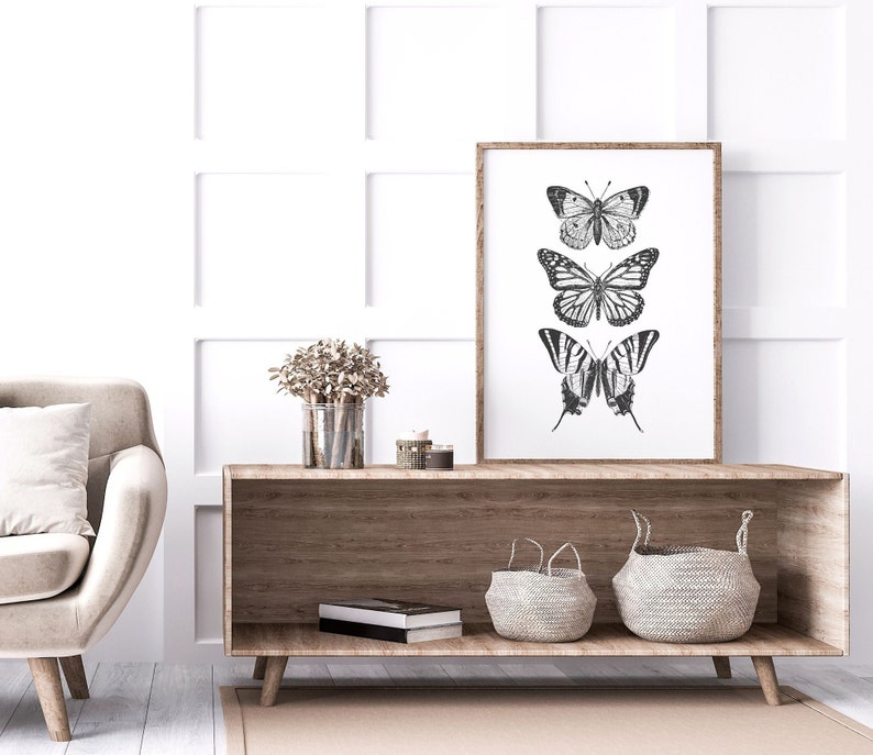 May include: A framed black and white print of three butterflies on a white background. The print is hanging on a white wall above a wooden shelf with two woven baskets and a book.