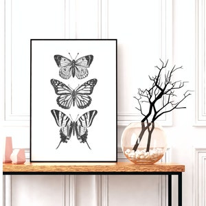 May include: Black and white print of three butterflies on a white background in a black frame. The butterflies are arranged vertically, with the largest butterfly at the bottom. The print is hanging on a wall above a wooden table with a vase of branches.