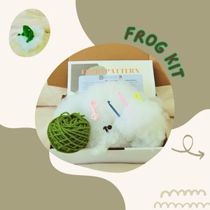 Könnte beinhalten: Ein Häkel-Set für eine Froschfigur. Das Set enthält einen Knäuel grüne Wolle, weiße Füllwatte, rosa und blaue Häkelnadeln und etwas rosa Garn. Die Box enthält auch eine Anleitung mit dem Text "FROG PATTERN".