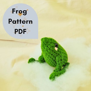 Op de afbeelding: Een handgemaakte groene gehaakte kikker met zwarte ogen en een kleine roze mond. De kikker zit op een bedje van witte pluisjes. Een lichtblauwe ovale afbeelding met de woorden "Frog Pattern PDF" staat op de achtergrond.