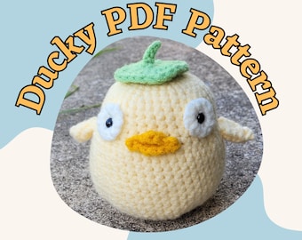May include: A crocheted yellow duck with white eyes, a yellow beak, and a green leaf on its head. The text "Ducky PDF Pattern" is in a curved, orange font around the duck.