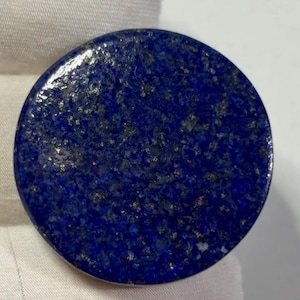Puede incluir: Un cabujón de piedra preciosa lapislázuli circular y pulido que presenta un color base azul real profundo con inclusiones de pirita de color dorado metálico dispersas por toda la superficie, creando un aspecto moteado y natural.
