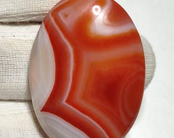 May include: Oval-shaped agate gemstone with a vibrant red and white banded pattern. The polished stone displays a natural, swirling design, showcasing the unique mineral formations. The gemstone is held against a white background.