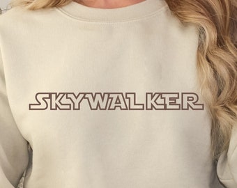 Może przedstawiać: Beżowa bluza z nadrukiem "SKYWALKER" w kolorze brązowym na przodzie. Bluzę nosi osoba z długimi blond włosami.