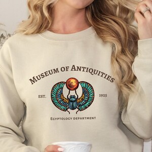 Puede incluir: Una sudadera beige con un estampado gr&aacute;fico de un escarabajo con alas y un disco solar. El texto "Museum of Antiquities Est. 1925 Egyptology Department" est&aacute; impreso debajo del gr&aacute;fico.