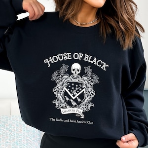 May include: Black sweatshirt featuring a white graphic of a coat of arms. The graphic includes a skull, a shield with stars and a sword, and three birds. The text "House of Black" and "Toujours Pur" are included in the graphic. The text "The Noble and Most Ancient Clan" is below the graphic.