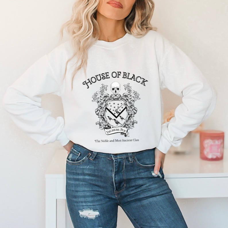 May include: White sweatshirt with a black and white graphic of a coat of arms with the text "House of Black" and "Toujours Pur" with the words "The Noble and Most Ancient Clan" below.