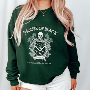 May include: A dark green sweatshirt with a white graphic design featuring a skull, a shield with a raven, and the text "House of Black, Toujours Pur, The Noble and Most Ancient Clan".