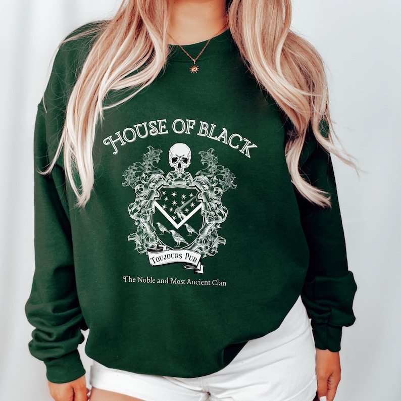 May include: A dark green sweatshirt with a white graphic design featuring a skull, a shield with a raven, and the text "House of Black, Toujours Pur, The Noble and Most Ancient Clan".