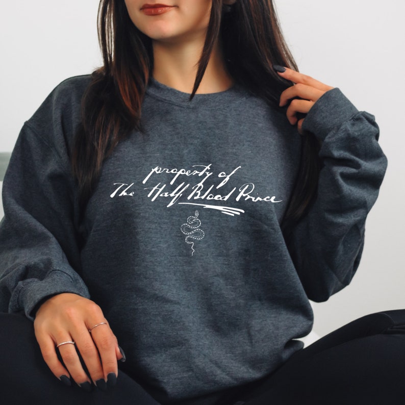 May include: A dark gray sweatshirt with a white graphic print that says "property of The Half Blood Prince" with a snake design below the text.