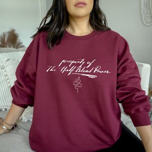 May include: Burgundy sweatshirt with white text that reads "property of The Half Blood Prince" with a snake design below the text.